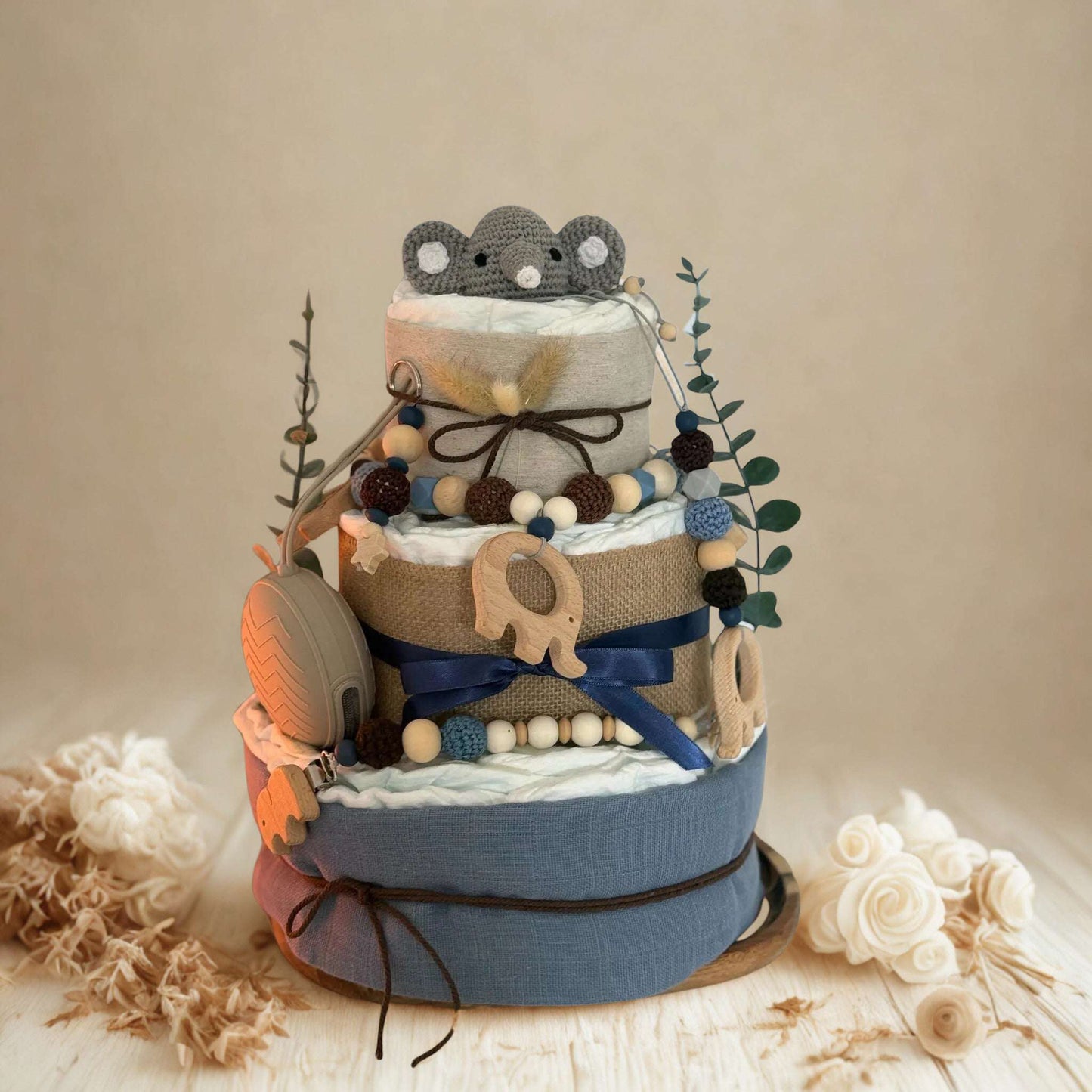 Blue and neutral safari diaper cake baby shower centerpiece featuring three tiers of rolled white diapers wrapped in soft blue fabric and natural burlap ribbon. Top tier decorated with a crocheted gray elephant plush, accented with dried pampas grass and eucalyptus greenery stems. Adorned with wooden elephant-shaped teething rings, silicone and wooden beaded pacifier clips in navy, brown, and cream tones, plus dark blue satin bow detail. Includes beige pacifier case attached to the side. Rustic boho newborn