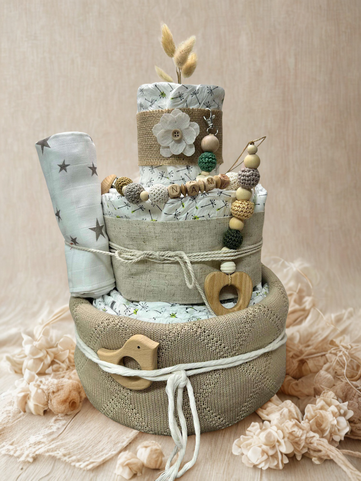 Neutral green and beige diaper cake baby shower centerpiece featuring three tiers of rolled white diapers wrapped in linen fabric and soft knit basket base. Decorated with wooden and silicone beaded pacifier clip personalized with letter beads spelling “NAME,” natural wood heart-shaped teething ring, wooden bird teether, and crochet beads in sage, cream, and mustard tones. Accented with burlap ribbon, fabric flower applique, dried bunny tail grass, and a rolled muslin blanket with gray star print. Rustic bo