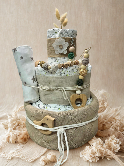 Neutral green and beige diaper cake baby shower centerpiece featuring three tiers of rolled white diapers wrapped in linen fabric and soft knit basket base. Decorated with wooden and silicone beaded pacifier clip personalized with letter beads spelling “NAME,” natural wood heart-shaped teething ring, wooden bird teether, and crochet beads in sage, cream, and mustard tones. Accented with burlap ribbon, fabric flower applique, dried bunny tail grass, and a rolled muslin blanket with gray star print. Rustic bo