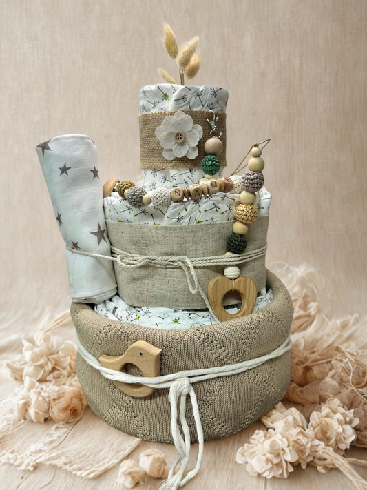 Neutral green and beige diaper cake baby shower centerpiece featuring three tiers of rolled white diapers wrapped in linen fabric and soft knit basket base. Decorated with wooden and silicone beaded pacifier clip personalized with letter beads spelling “NAME,” natural wood heart-shaped teething ring, wooden bird teether, and crochet beads in sage, cream, and mustard tones. Accented with burlap ribbon, fabric flower applique, dried bunny tail grass, and a rolled muslin blanket with gray star print. Rustic bo