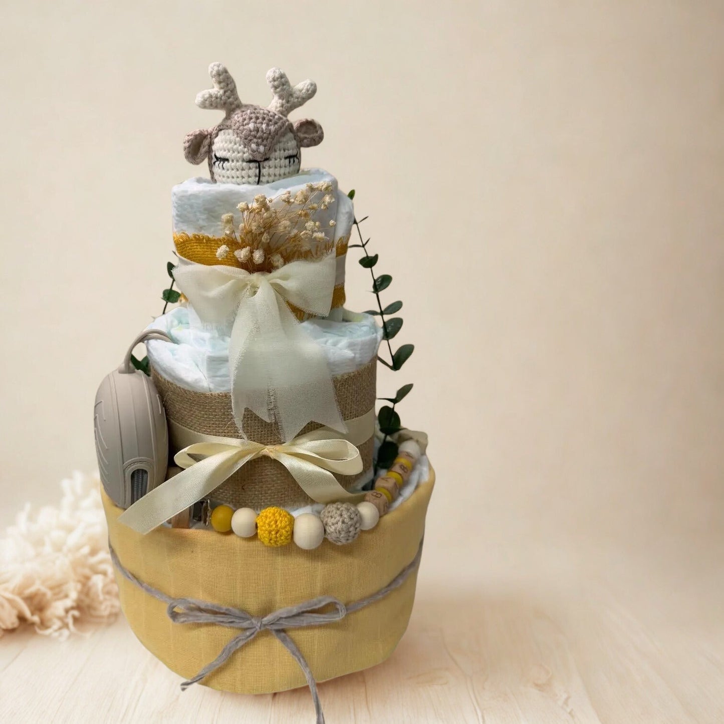 Safari giraffe diaper cake baby shower centerpiece designed in soft yellow, cream, and natural burlap tones. This three-tier arrangement features rolled white diapers wrapped in warm pastel yellow fabric at the base and finished with a delicate gray twine bow for a charming rustic touch.

The middle tier is wrapped in natural burlap ribbon and tied with an elegant ivory satin bow, complemented by a coordinating silicone and crochet beaded pacifier clip in mustard, cream, and soft gray hues. A matching neutr