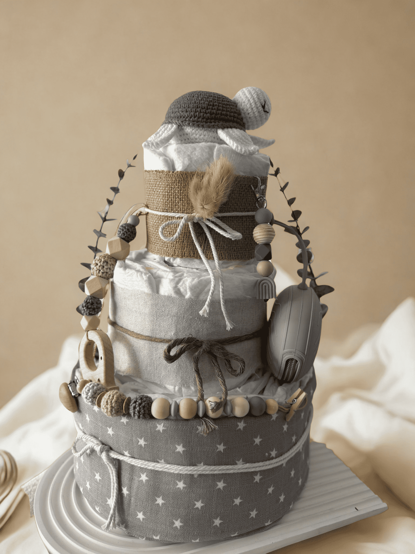 Neutral teddy bear diaper cake baby shower centerpiece featuring three tiers of rolled white diapers wrapped in natural burlap and soft gray star-print fabric. Top tier decorated with a crocheted teddy bear plush wearing a matching hat, accented with dried bunny tail grass and rustic twine bow. Adorned with silicone and crochet beaded pacifier clips in beige, gray, and cream tones, wooden bead garlands, and natural wood teething rings including animal-shaped designs. Includes a soft knitted baby bonnet and 