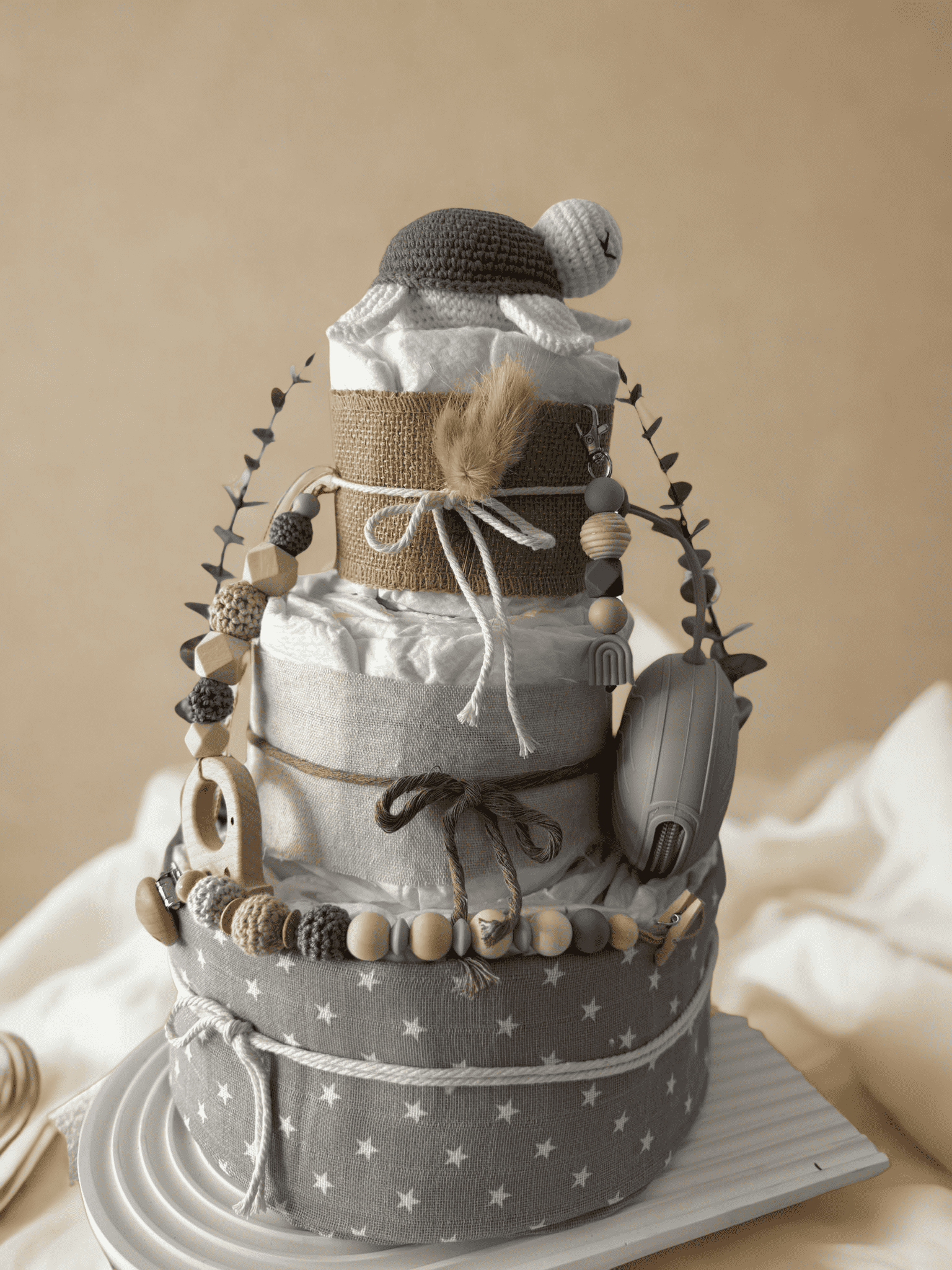 Neutral teddy bear diaper cake baby shower centerpiece featuring three tiers of rolled white diapers wrapped in natural burlap and soft gray star-print fabric. Top tier decorated with a crocheted teddy bear plush wearing a matching hat, accented with dried bunny tail grass and rustic twine bow. Adorned with silicone and crochet beaded pacifier clips in beige, gray, and cream tones, wooden bead garlands, and natural wood teething rings including animal-shaped designs. Includes a soft knitted baby bonnet and 