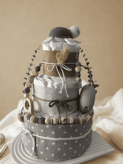 Neutral teddy bear diaper cake baby shower centerpiece featuring three tiers of rolled white diapers wrapped in natural burlap and soft gray star-print fabric. Top tier decorated with a crocheted teddy bear plush wearing a matching hat, accented with dried bunny tail grass and rustic twine bow. Adorned with silicone and crochet beaded pacifier clips in beige, gray, and cream tones, wooden bead garlands, and natural wood teething rings including animal-shaped designs. Includes a soft knitted baby bonnet and 