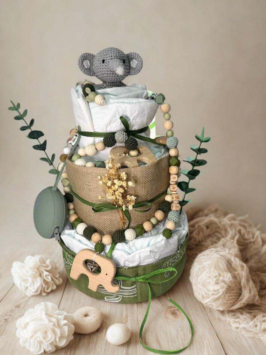 Green cactus-themed diaper cake baby shower centerpiece featuring three tiers of rolled white diapers wrapped in sage and olive green fabric with burlap trim and satin ribbon bows. Top tier decorated with a crocheted gray elephant plush, accented with dried wheat grass and eucalyptus greenery. Adorned with wooden and silicone beaded pacifier clip in neutral green, cream, and gray tones, plus natural wood teething clip detail. Includes matching green pacifier case attached to the side. Base tier wrapped in o
