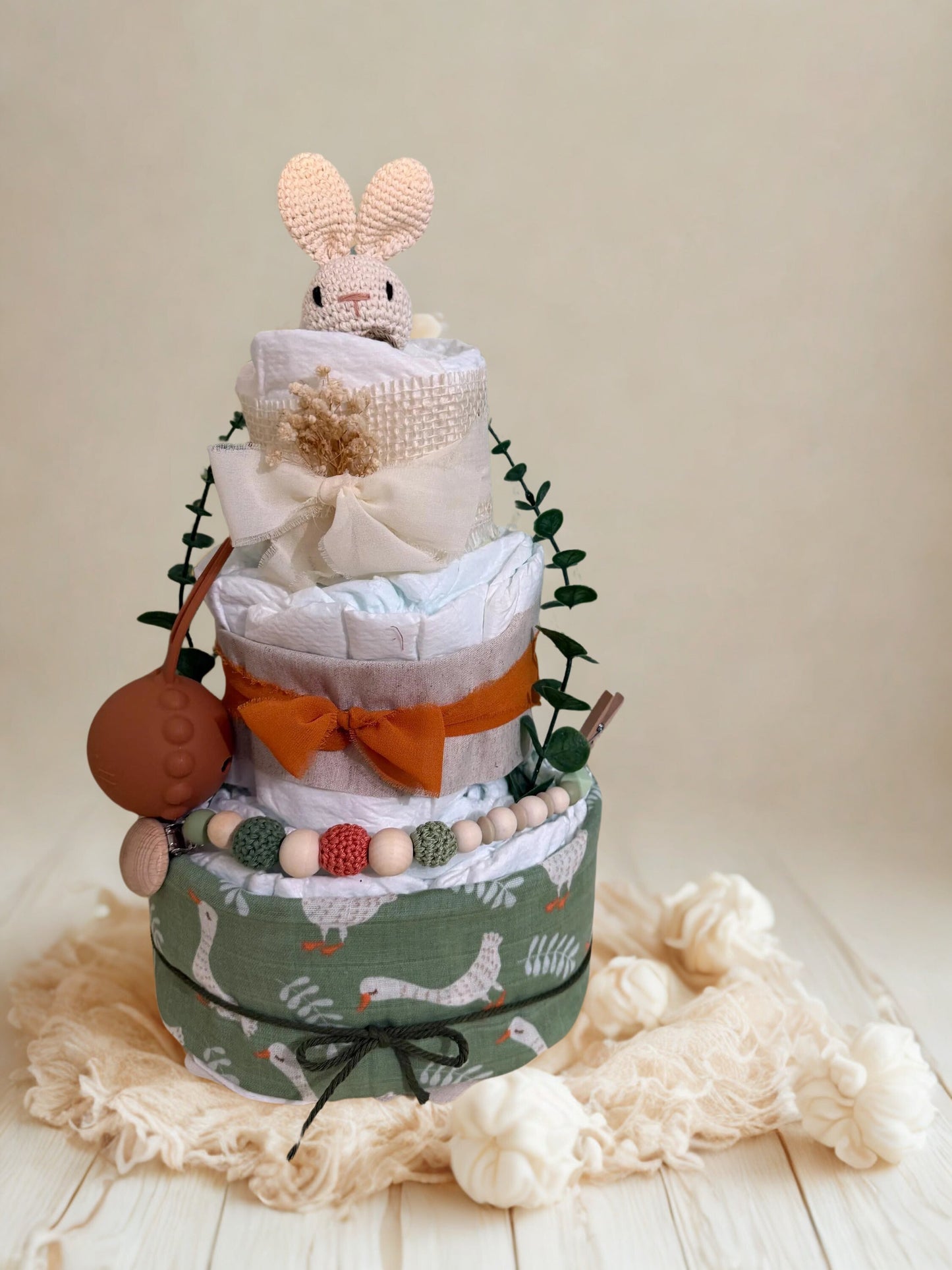 Rustic woodland bunny diaper cake baby shower centerpiece featuring three tiers of rolled white diapers in warm autumn-inspired tones. The base tier is wrapped in sage green fabric with a charming white goose print and tied with dark twine for a cozy farmhouse finish.

The middle tier is accented with soft linen fabric and a burnt orange chiffon bow, paired with a coordinating silicone and crochet beaded pacifier clip in earthy shades of olive, terracotta, cream, and natural wood. A matching warm brown paci