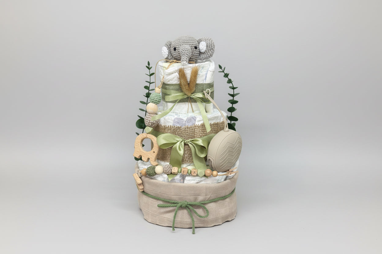 Green safari diaper cake baby shower centerpiece featuring three tiers of rolled white diapers wrapped in burlap ribbon and sage green satin bows. Top tier decorated with a crocheted gray elephant plush, accented with dried pampas grass and eucalyptus greenery. Adorned with wooden elephant-shaped teething ring, silicone and crochet beaded pacifier clip in green, cream, and natural wood tones, personalized with wooden letter beads spelling “LEA.” Includes beige pacifier case attached to the side. Rustic boho