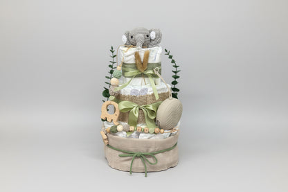 Green safari diaper cake baby shower centerpiece featuring three tiers of rolled white diapers wrapped in burlap ribbon and sage green satin bows. Top tier decorated with a crocheted gray elephant plush, accented with dried pampas grass and eucalyptus greenery. Adorned with wooden elephant-shaped teething ring, silicone and crochet beaded pacifier clip in green, cream, and natural wood tones, personalized with wooden letter beads spelling “LEA.” Includes beige pacifier case attached to the side. Rustic boho