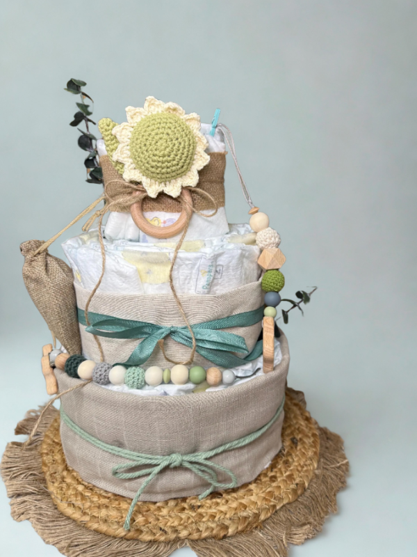 Sunflower-themed diaper cake baby shower centerpiece featuring three tiers of rolled white diapers wrapped in natural linen and burlap ribbon with sage green satin bows. Top tier decorated with a crocheted sunflower plush in soft yellow and light green tones, accented with eucalyptus greenery and rustic twine detail. Adorned with wooden teething ring, silicone and crochet beaded pacifier clip in green, cream, and gray shades, and neutral wooden bead garland. Elegant boho newborn gift arrangement displayed o