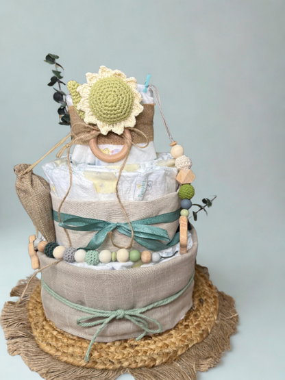 Sunflower-themed diaper cake baby shower centerpiece featuring three tiers of rolled white diapers wrapped in natural linen and burlap ribbon with sage green satin bows. Top tier decorated with a crocheted sunflower plush in soft yellow and light green tones, accented with eucalyptus greenery and rustic twine detail. Adorned with wooden teething ring, silicone and crochet beaded pacifier clip in green, cream, and gray shades, and neutral wooden bead garland. Elegant boho newborn gift arrangement displayed o
