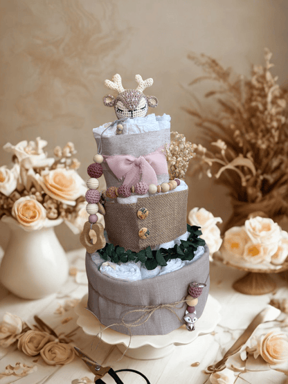 Elegant neutral diaper cake baby shower centerpiece featuring three tiers of rolled white diapers wrapped in soft gray linen and burlap fabric. Top tier decorated with a crocheted deer plush in beige and cream tones, accented with a blush pink bow. Adorned with wooden and silicone beaded pacifier clip in mauve and natural wood shades, wooden teething ring, dried flowers, and eucalyptus greenery for a rustic woodland theme. Styled on a white cake stand with soft beige floral background, roses, and pampas gra