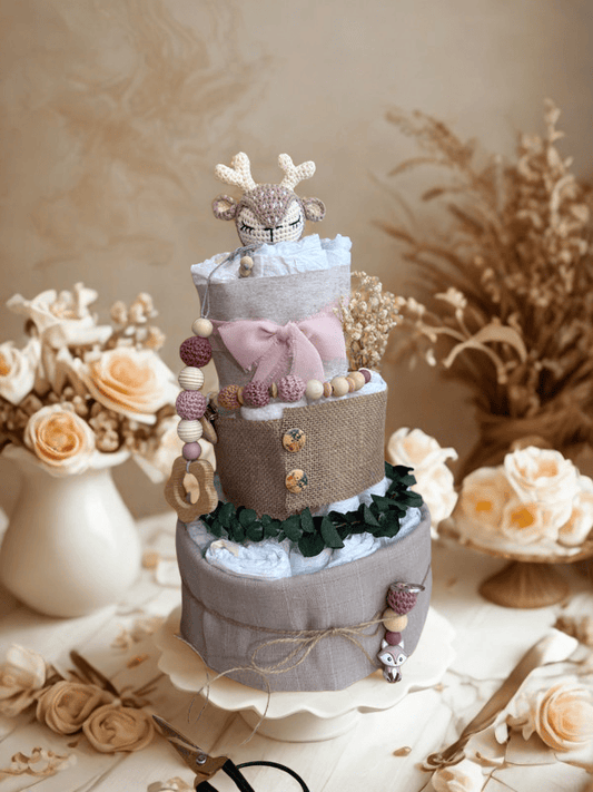 Elegant neutral diaper cake baby shower centerpiece featuring three tiers of rolled white diapers wrapped in soft gray linen and burlap fabric. Top tier decorated with a crocheted deer plush in beige and cream tones, accented with a blush pink bow. Adorned with wooden and silicone beaded pacifier clip in mauve and natural wood shades, wooden teething ring, dried flowers, and eucalyptus greenery for a rustic woodland theme. Styled on a white cake stand with soft beige floral background, roses, and pampas gra