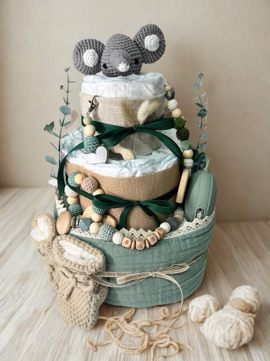 Green and neutral diaper cake baby shower centerpiece featuring three tiers of rolled white diapers wrapped in sage green muslin and burlap ribbon. Top tier adorned with a crocheted gray elephant plush toy, accented with eucalyptus stems and dried grass detail. Decorated with wooden and silicone beaded pacifier clip personalized with letter beads spelling “BABY,” wooden teething ring, lace trim, and dark green satin bows. Includes matching green pacifier case attached to the side. Rustic boho nursery gift s