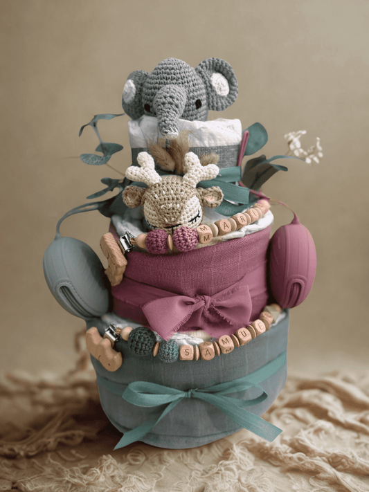 Pink and teal woodland diaper cake baby shower centerpiece featuring three tiers of rolled white diapers wrapped in dusty rose and sage green fabric with soft ribbon bows. Top tier decorated with a crocheted gray elephant plush, accented with eucalyptus greenery and dried grass detail. Middle tier adorned with a crocheted beige deer plush and silicone beaded pacifier clip personalized with wooden letter beads spelling “MARA.” Bottom tier features additional beaded clip spelling “SAMUEL,” wooden animal-shape