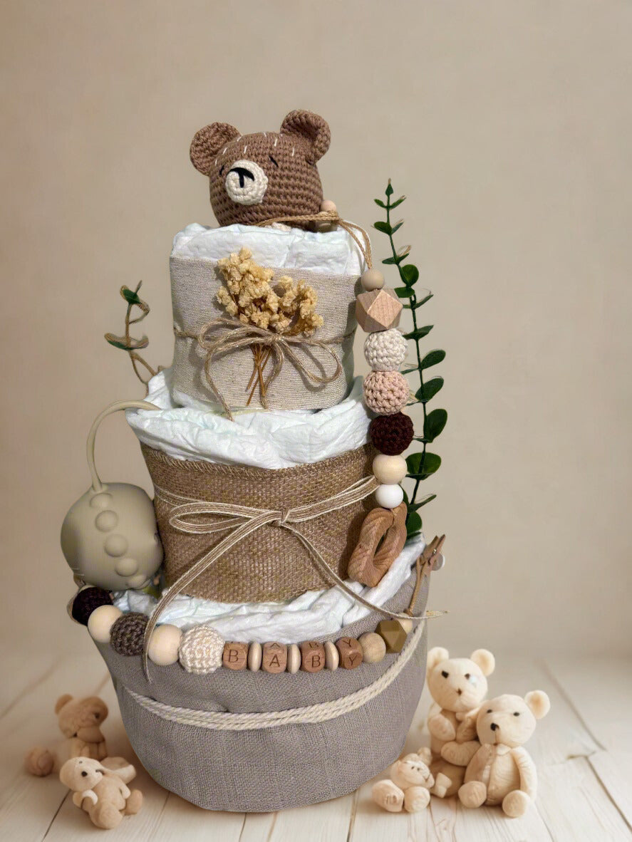Neutral teddy bear diaper cake baby shower centerpiece featuring three tiers of rolled white diapers in soft beige, taupe, and natural linen textures. The base tier is wrapped in warm gray fabric and finished with a rustic rope accent for a cozy, modern touch.

The middle tier is adorned with burlap-style fabric and tied with delicate twine, complemented by a silicone and crochet beaded pacifier clip in earthy tones of cream, mocha, blush, and walnut. Wooden letter beads spelling “BABY” add a charming perso