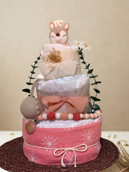 Blush pink deer diaper cake baby shower centerpiece featuring three tiers of rolled white diapers wrapped in soft tulle, cream fabric, and a coral pink sun-print band. Top tier decorated with a crocheted fawn plush in warm beige and blush tones, accented with delicate dried florals and a sheer ivory bow for a romantic boho touch.

Middle tier finished with a dusty rose ribbon bow and adorned with a coordinating silicone and crochet beaded pacifier clip in shades of blush, peach, and cream. Includes a matchi