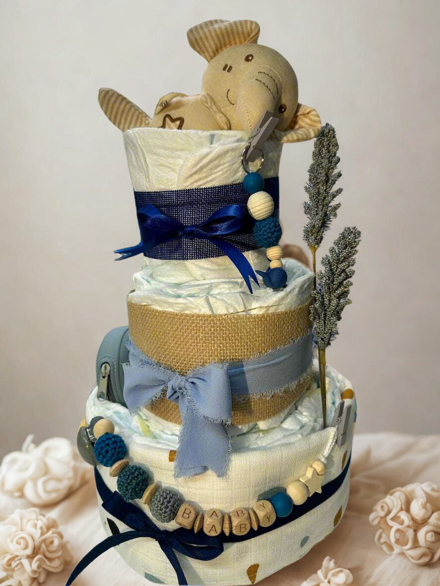 Three-tier blue safari diaper cake featuring rolled white diapers wrapped in burlap, soft blue fabric, and patterned cream material with subtle accents. The base tier is tied with a navy ribbon and decorated with a personalized wooden and silicone beaded pacifier clip spelling “BABY.”

The middle tier is wrapped in natural burlap and finished with a soft dusty blue bow, adding a rustic yet elegant touch. The top tier is secured with a navy ribbon and topped with a plush beige elephant toy peeking out from t