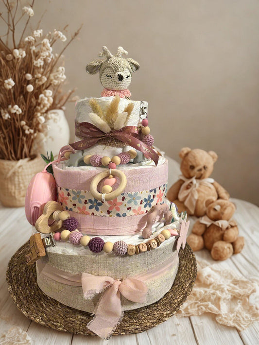 Pink floral diaper cake baby shower centerpiece featuring three tiers of rolled white diapers wrapped in soft blush and beige fabric with delicate flower print trim. Top tier decorated with a crocheted deer plush in cream and pastel pink tones, accented with dried pampas grass and satin ribbon bow. Adorned with wooden and silicone beaded pacifier clip personalized with letter beads spelling “BABY,” natural wood teething ring, butterfly charm, and pink pacifier case attached to the side. Rustic boho newborn 