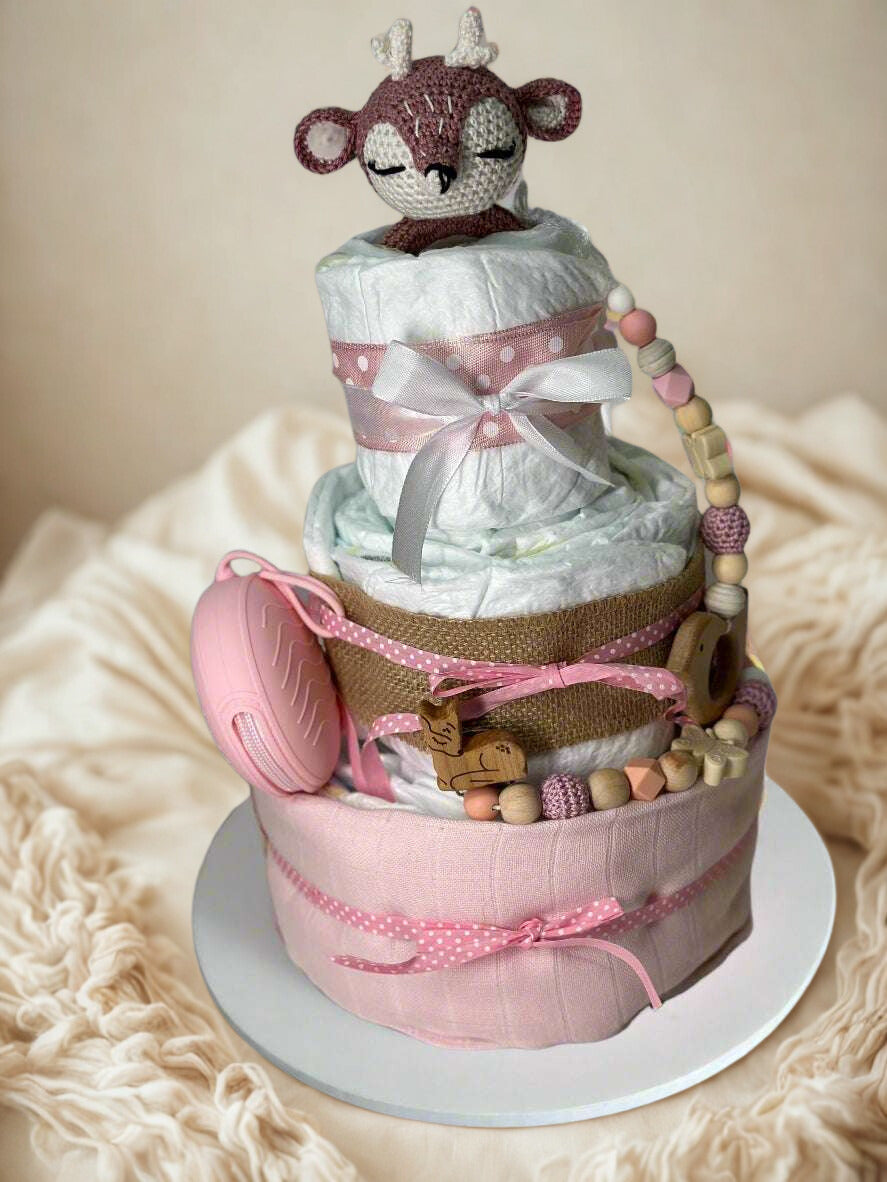 Three-tier pink woodland diaper cake featuring rolled white diapers wrapped in soft blush fabric, burlap, and pink polka dot ribbon bows. A crocheted fawn plush with closed eyes sits on the top tier, adding a sweet forest-inspired touch. The cake is decorated with a pink silicone pacifier case, wooden deer teether, and a coordinating silicone and wooden beaded pacifier clip in blush, cream, and natural tones, creating a charming baby girl shower centerpiece.
