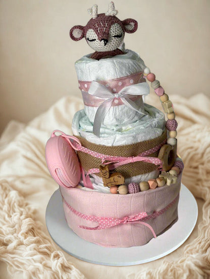 Three-tier pink woodland diaper cake featuring rolled white diapers wrapped in soft blush fabric, burlap, and pink polka dot ribbon bows. A crocheted fawn plush with closed eyes sits on the top tier, adding a sweet forest-inspired touch. The cake is decorated with a pink silicone pacifier case, wooden deer teether, and a coordinating silicone and wooden beaded pacifier clip in blush, cream, and natural tones, creating a charming baby girl shower centerpiece.
