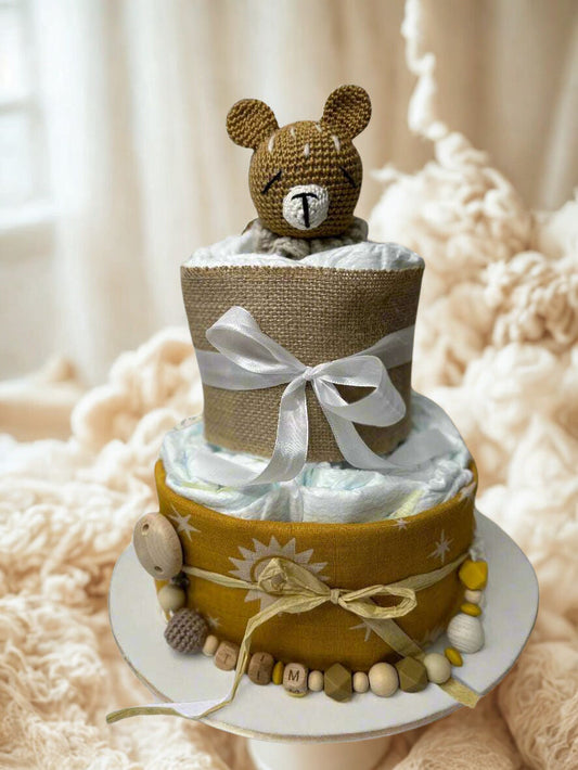 Mustard and neutral teddy bear diaper cake baby shower centerpiece featuring two tiers of rolled white diapers styled in warm golden and natural burlap tones. The base tier is wrapped in mustard fabric with subtle sun and star prints, tied with rustic twine for a cozy, earthy finish.

The top tier is covered in textured burlap and finished with a soft white satin ribbon bow, creating a beautiful contrast of rustic and elegant details. A handmade crocheted teddy bear in warm caramel brown crowns the design, 