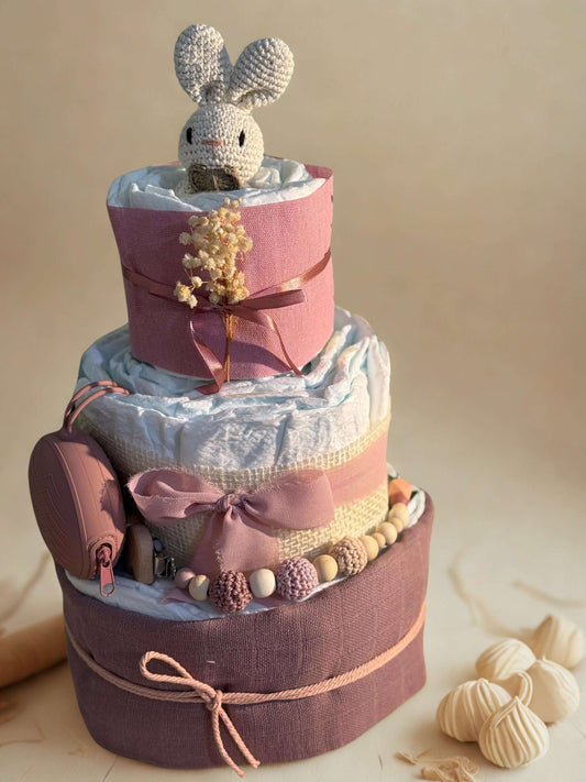 Blush pink bunny diaper cake baby shower centerpiece featuring three tiers of rolled white diapers wrapped in soft mauve and dusty rose fabrics. The bottom tier is finished with delicate twine, creating a warm rustic foundation in elegant muted tones.

The middle tier is accented with natural burlap ribbon and a soft rose chiffon bow, paired with a coordinating silicone and crochet beaded pacifier clip in shades of blush, cream, and nude. A matching rose pink pacifier case is attached to the side for a prac
