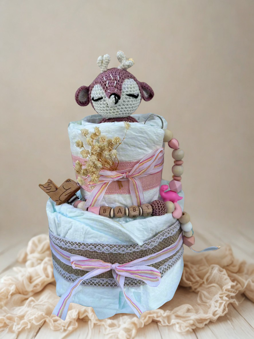 Pink and pastel diaper cake baby shower centerpiece featuring two tiers of rolled white diapers wrapped in soft blue tissue and burlap lace trim. Top tier decorated with a crocheted pink and cream deer plush toy, accented with dried baby’s breath flowers and a striped pink ribbon bow. Adorned with a wooden bird pacifier clip, silicone and wooden beaded teether personalized with letter beads spelling “BABY,” and a pink flamingo charm. Rustic boho newborn gift arrangement styled with lace details, natural tex