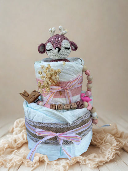 Pink and pastel diaper cake baby shower centerpiece featuring two tiers of rolled white diapers wrapped in soft blue tissue and burlap lace trim. Top tier decorated with a crocheted pink and cream deer plush toy, accented with dried baby’s breath flowers and a striped pink ribbon bow. Adorned with a wooden bird pacifier clip, silicone and wooden beaded teether personalized with letter beads spelling “BABY,” and a pink flamingo charm. Rustic boho newborn gift arrangement styled with lace details, natural tex