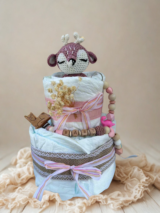 Pink and pastel diaper cake baby shower centerpiece featuring two tiers of rolled white diapers wrapped in soft blue tissue and burlap lace trim. Top tier decorated with a crocheted pink and cream deer plush toy, accented with dried baby’s breath flowers and a striped pink ribbon bow. Adorned with a wooden bird pacifier clip, silicone and wooden beaded teether personalized with letter beads spelling “BABY,” and a pink flamingo charm. Rustic boho newborn gift arrangement styled with lace details, natural tex