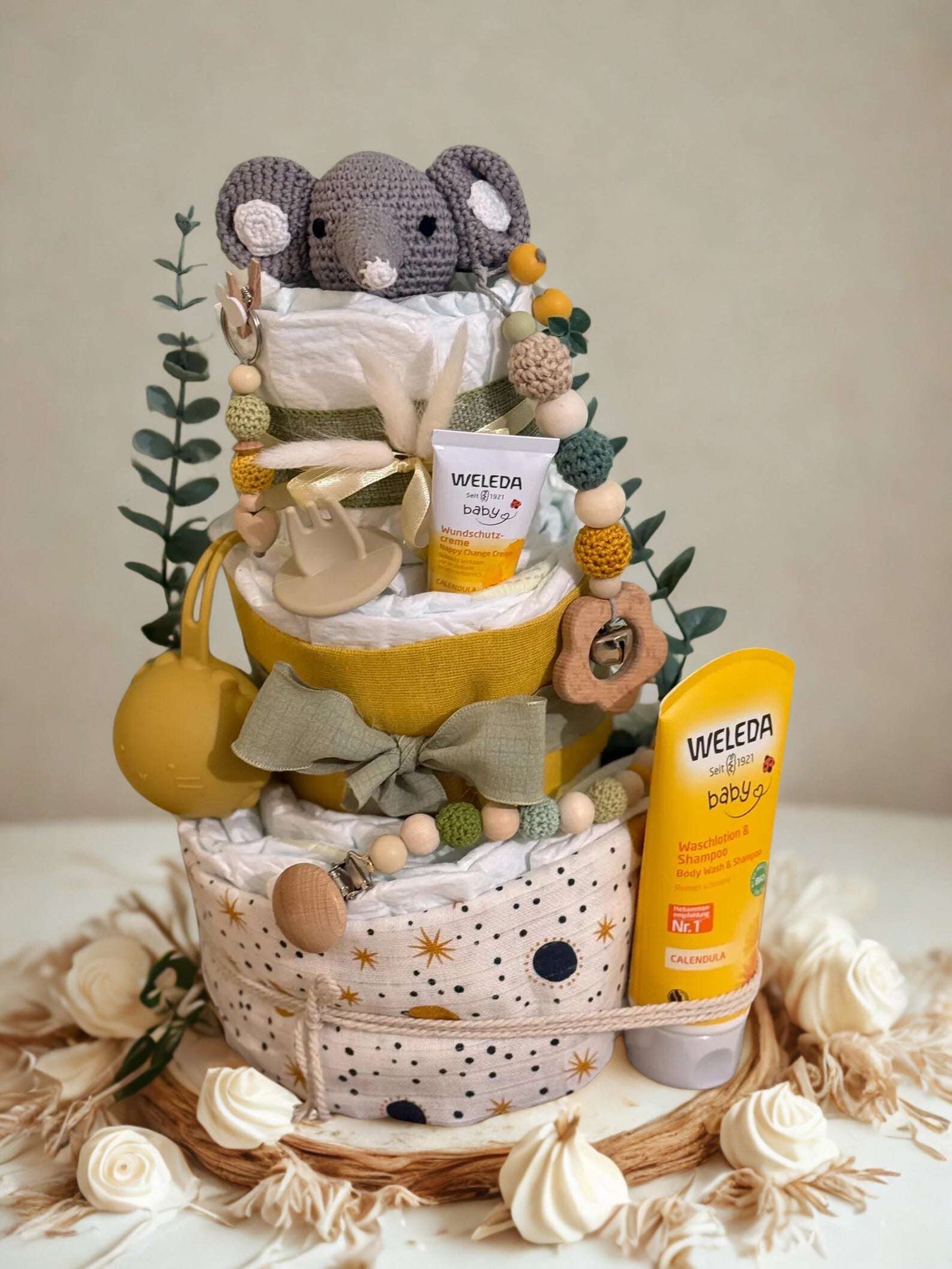 Neutral yellow and green diaper cake baby shower gift featuring three tiers of rolled white diapers wrapped in mustard yellow and sage green fabric. Top tier decorated with a crocheted gray elephant plush, eucalyptus greenery, and a beaded pacifier clip in earthy tones. Includes visible baby skincare products labeled “WELEDA baby,” with Calendula diaper cream and baby wash shampoo displayed on and beside the cake. Rustic boho nursery centerpiece styled with wooden teething toys, silicone beads, star-pattern