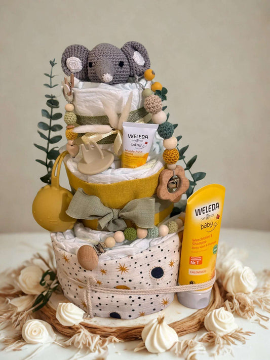 Neutral yellow and green diaper cake baby shower gift featuring three tiers of rolled white diapers wrapped in mustard yellow and sage green fabric. Top tier decorated with a crocheted gray elephant plush, eucalyptus greenery, and a beaded pacifier clip in earthy tones. Includes visible baby skincare products labeled “WELEDA baby,” with Calendula diaper cream and baby wash shampoo displayed on and beside the cake. Rustic boho nursery centerpiece styled with wooden teething toys, silicone beads, star-pattern