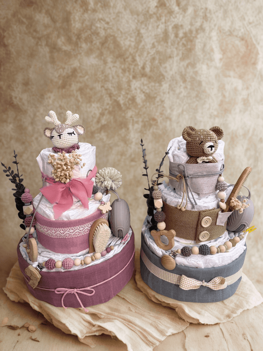 Two handcrafted diaper cake baby shower centerpieces displayed side by side on a neutral backdrop, featuring three-tier stacks of rolled white diapers wrapped in soft fabric. Left diaper cake in dusty rose and blush tones with lace trim, pink bow, wooden teething ring, silicone bead pacifier clip, baby brush, dried flowers, and crocheted deer plush topper. Right diaper cake in gray and beige rustic style with burlap ribbon, gingham bow, wooden buttons, wooden teether, beaded pacifier chain, baby brush, and 