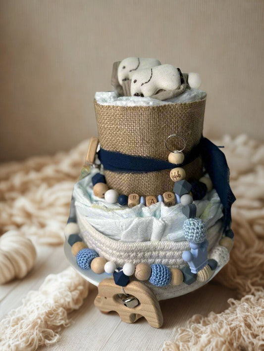 Navy and neutral baby boy diaper cake centerpiece featuring two beautifully styled tiers of rolled white diapers in soft blue and natural textures. The base tier is wrapped in cream knit trim and accented with a wooden elephant teether, adding a sweet safari-inspired touch.

The middle tier is decorated with a coordinating silicone and crochet beaded pacifier clip in shades of navy, powder blue, white, and natural wood. Wooden letter beads spelling “BABY” add a charming detail, while a personalized wooden b
