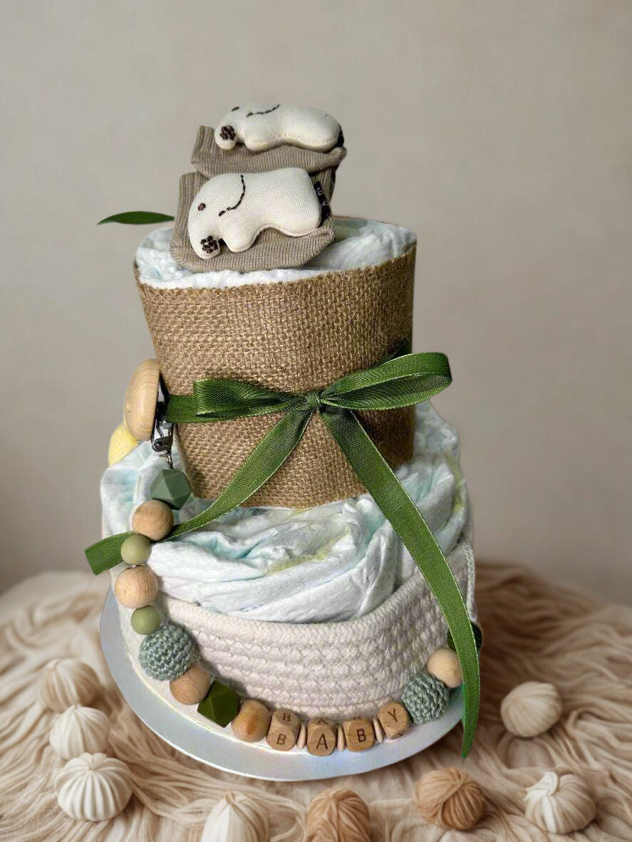 Neutral sage baby diaper cake centerpiece featuring two tiers of rolled white diapers styled in soft green and natural burlap textures. The base tier is wrapped in cozy cream knit trim, creating a warm and timeless foundation, and finished with wooden letter beads spelling “BABY” for a sweet decorative touch.

The top tier is wrapped in rustic burlap and tied with a muted olive green satin ribbon, adding an elegant woodland accent. Adorable baby socks styled as a tiny animal face crown the top, making this 