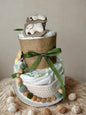 Neutral sage baby diaper cake centerpiece featuring two tiers of rolled white diapers styled in soft green and natural burlap textures. The base tier is wrapped in cozy cream knit trim, creating a warm and timeless foundation, and finished with wooden letter beads spelling “BABY” for a sweet decorative touch.

The top tier is wrapped in rustic burlap and tied with a muted olive green satin ribbon, adding an elegant woodland accent. Adorable baby socks styled as a tiny animal face crown the top, making this 