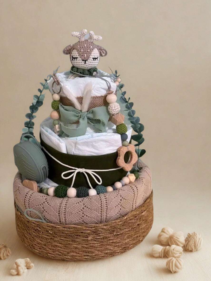 Woodland deer diaper cake baby shower centerpiece featuring three tiers of rolled white diapers wrapped in rustic burlap, soft sage ribbon, and a contrasting dark accent band with twine bow detail. Top tier decorated with a crocheted beige deer plush with delicate antlers, adding a charming forest-inspired touch.

Accented with eucalyptus greenery garland and silicone and crochet beaded pacifier clips in sage, forest green, cream, and natural wood tones. Includes a coordinating sage green pacifier case atta
