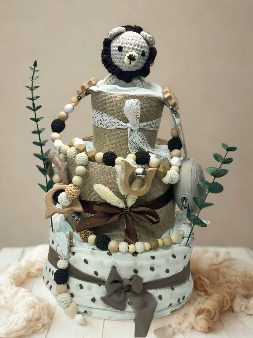Three-tier neutral safari diaper cake featuring rolled white diapers wrapped in burlap and soft polka dot fabric in cream and taupe tones. The base tier is tied with a mocha ribbon bow, while the middle tier is accented with a rich brown satin ribbon and dried floral details for a rustic touch.

The top tier is finished with delicate lace trim and crowned with a handmade crocheted lion plush in soft cream with a dark brown mane. The cake is decorated with coordinating wooden and silicone beaded pacifier cli