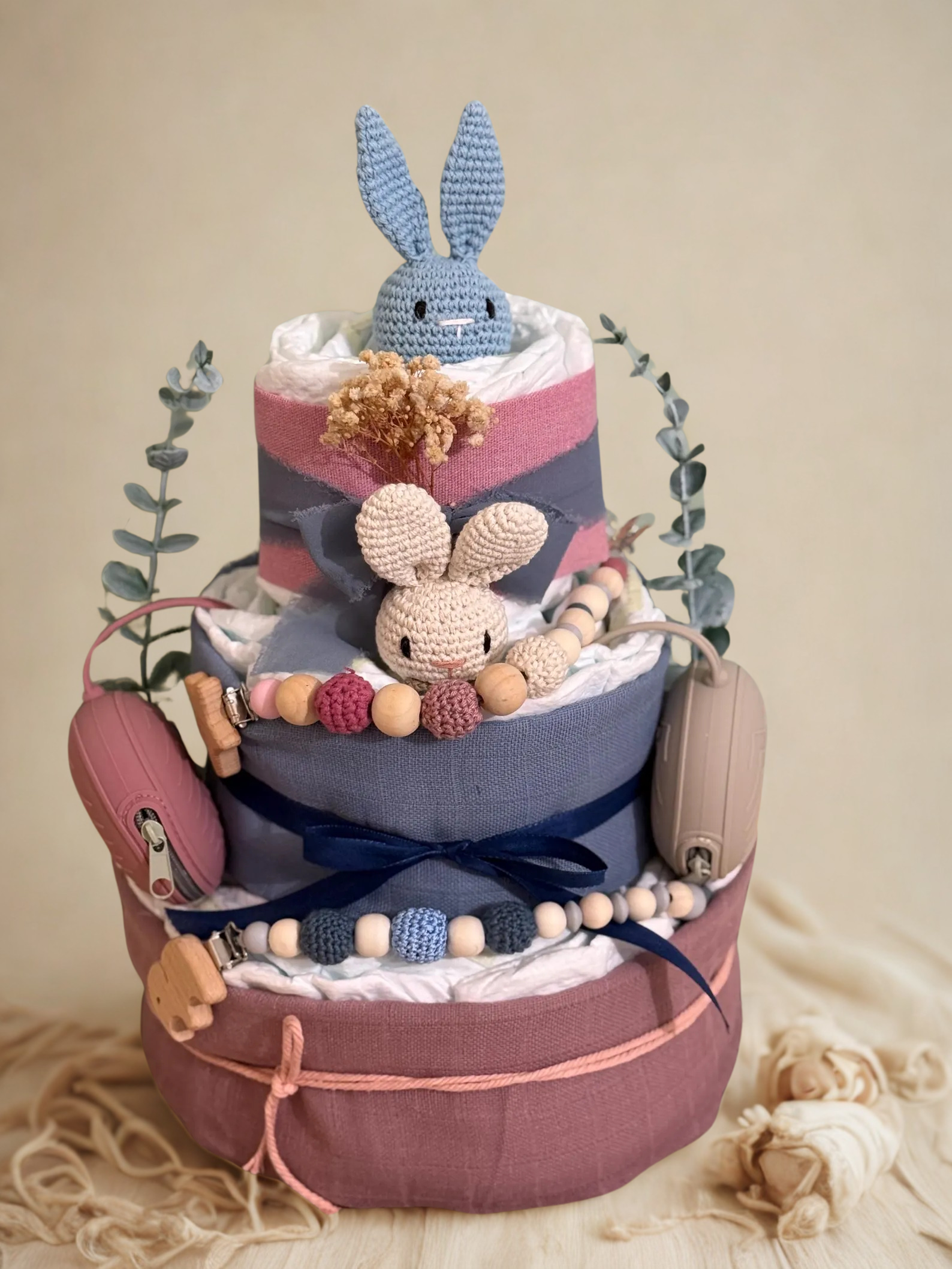 Dusty rose and slate blue bunny diaper cake baby shower centerpiece featuring three tiers of rolled white diapers wrapped in soft mauve, blush, and denim blue fabrics. The base tier is finished with delicate pink twine, creating a romantic yet modern boho foundation.

The middle tier is wrapped in muted blue fabric and accented with a navy satin bow, complemented by coordinating silicone and crochet beaded pacifier clips in shades of blush, cream, rose, and blue. Matching blush and taupe pacifier cases are 