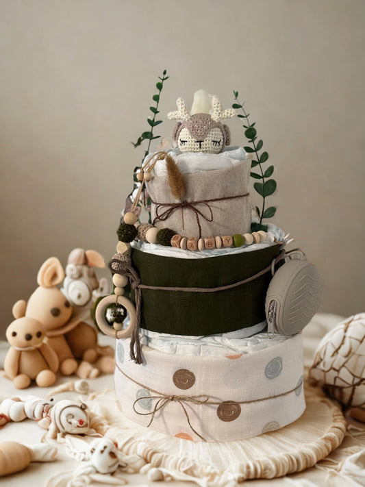 Green and neutral woodland diaper cake baby shower gift featuring three tiers of rolled white diapers wrapped in sage green and beige fabric with polka dot accents. Top tier decorated with a crocheted deer plush in cream and taupe tones, surrounded by eucalyptus greenery and natural twine bow. Adorned with wooden teething ring, silicone and wooden beaded pacifier clip personalized with letter beads spelling “BABY,” and a gray pacifier case attached to the side. Rustic boho nursery centerpiece styled with ea