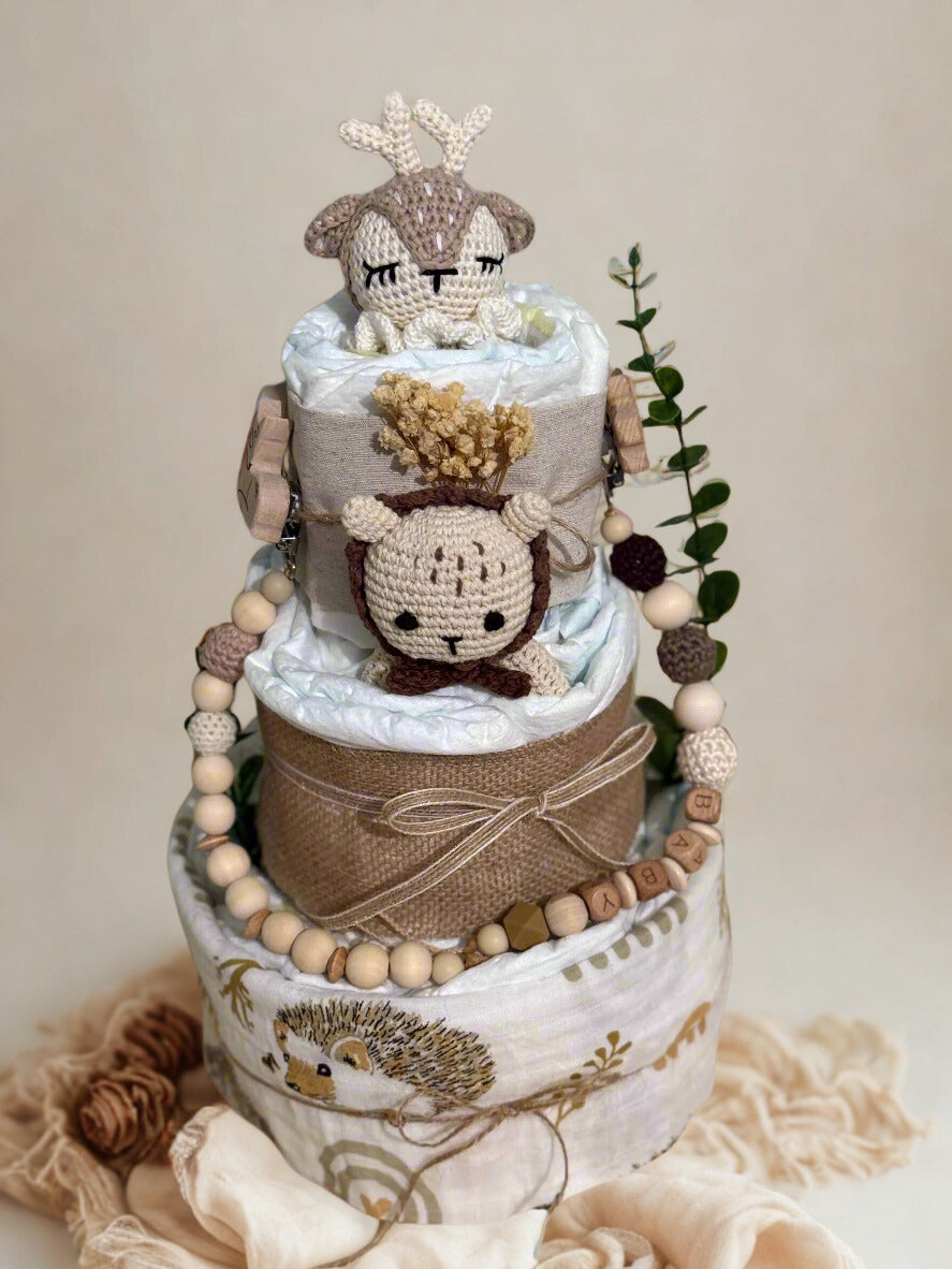 Woodland animal diaper cake baby shower centerpiece featuring three tiers of rolled white diapers wrapped in natural burlap and soft neutral fabric with hedgehog print on the base tier. Top decorated with crocheted beige and cream deer plush, with matching crocheted lion plush on the middle tier. Accented with dried flowers, eucalyptus greenery, and rustic twine bows for a boho nursery theme. Adorned with wooden and silicone beaded pacifier clip personalized with letter beads spelling “BABY,” natural wood t