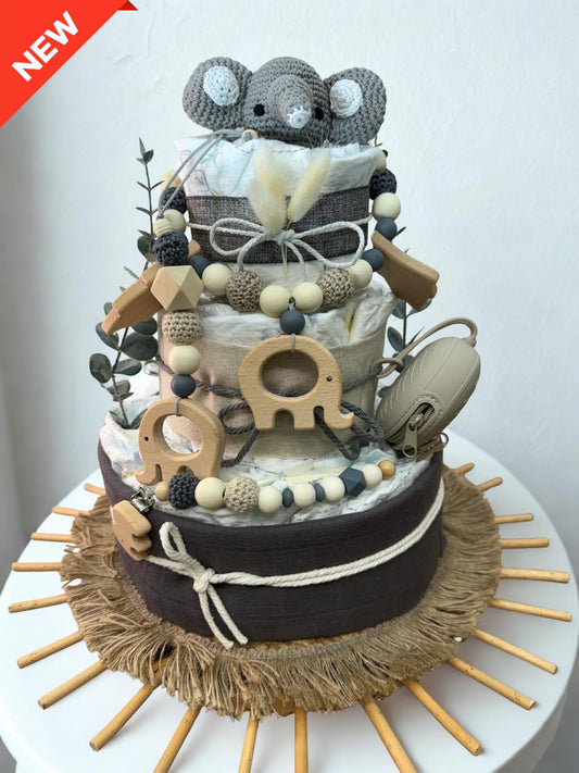 Neutral safari diaper cake baby shower centerpiece featuring three tiers of rolled white diapers wrapped in beige and charcoal fabric with rustic twine bows. Top tier decorated with a crocheted gray elephant plush, accented with dried pampas grass and eucalyptus greenery. Adorned with wooden elephant-shaped teething rings, geometric wooden beads, and silicone beaded pacifier clips in cream, taupe, and dark gray tones. Includes a beige pacifier case attached to the side. Rustic boho newborn gift arrangement 