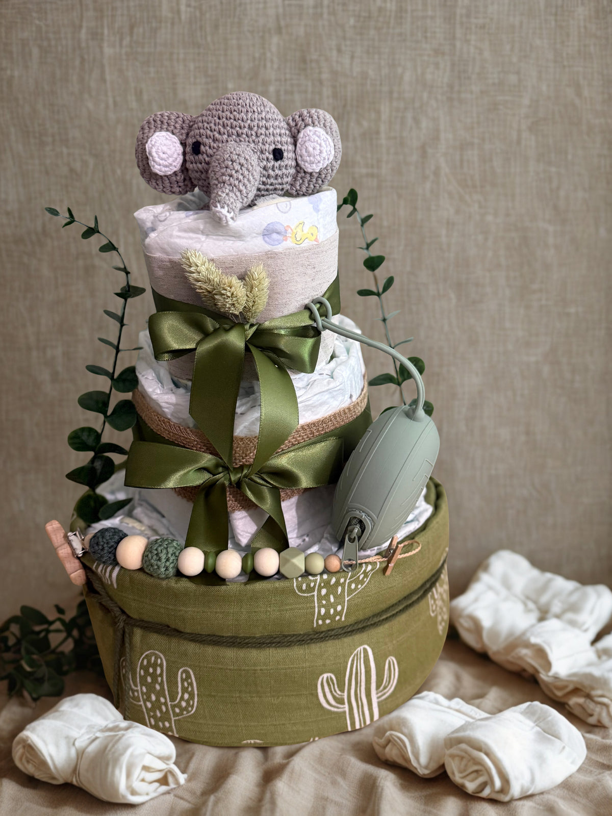 Green cactus-themed diaper cake baby shower centerpiece featuring three tiers of rolled white diapers wrapped in sage and olive green fabric with burlap trim and satin ribbon bows. Top tier decorated with a crocheted gray elephant plush, accented with dried wheat grass and eucalyptus greenery. Adorned with wooden and silicone beaded pacifier clip in neutral green, cream, and gray tones, plus natural wood teething clip detail. Includes matching green pacifier case attached to the side. Base tier wrapped in o