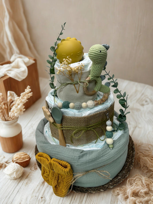 Sage green dinosaur diaper cake baby shower centerpiece featuring three tiers of rolled white diapers styled in soft mint, olive, and warm mustard tones. The base tier is wrapped in airy sage muslin fabric and finished with rustic twine, accented by a cozy mustard knit baby sweater for an extra thoughtful touch.

The middle tier is trimmed in natural burlap and tied with moss green cord, complemented by a silicone and crochet beaded pacifier clip in shades of eucalyptus, cream, and muted teal. A coordinatin
