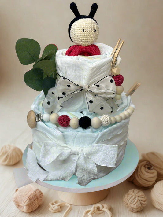 Ladybug diaper cake baby shower centerpiece featuring two tiers of rolled white diapers styled in crisp white with playful red and black accents. The base tier is wrapped in soft white fabric and finished with a classic white bow, creating a clean and elegant foundation.

The top tier is adorned with a sheer white ribbon with black polka dots, perfectly matching the charming handmade crocheted ladybug doll crowned at the top. The doll features delicate black antennae, a cream face, and a bright red body for