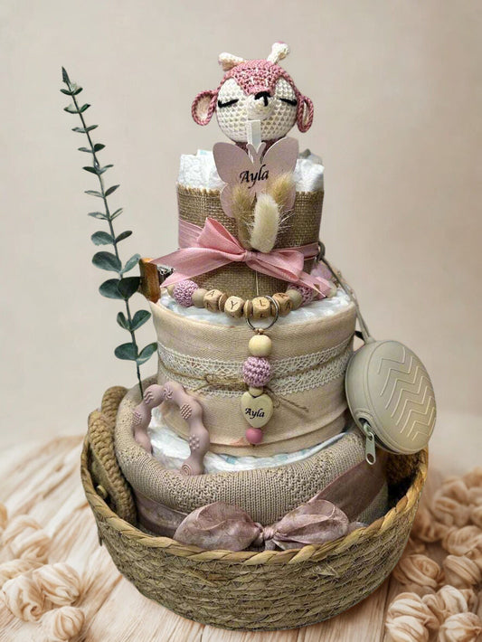 Three-tier blush and neutral woodland diaper cake arranged in a woven basket, featuring rolled white diapers wrapped in soft beige, burlap, and dusty pink fabrics with lace and ribbon accents. A crocheted fawn plush with pink details sits on the top tier beside a butterfly-shaped name topper reading “Ayla.”

The cake is decorated with a personalized wooden and silicone beaded pacifier clip with matching blush and cream tones, a coordinating teething ring, and a beige silicone pacifier case attached at the s