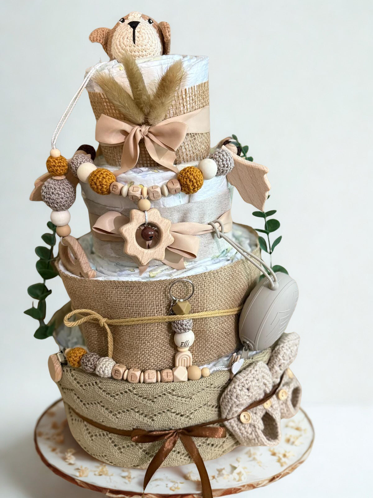 Neutral boho diaper cake baby shower gift featuring three tiers of rolled white diapers wrapped in burlap, knit fabric, and soft beige ribbon bows. Top tier decorated with a crocheted brown and cream puppy plush, accented with pampas grass detail. Adorned with wooden and silicone beaded pacifier clips personalized with letter beads spelling “ELLI,” natural wood teething ring, sun-shaped wooden rattle, and eucalyptus greenery stems. Includes beige pacifier case attached to the side and cozy knitted baby boot