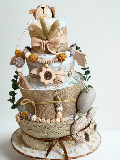 Neutral boho diaper cake baby shower gift featuring three tiers of rolled white diapers wrapped in burlap, knit fabric, and soft beige ribbon bows. Top tier decorated with a crocheted brown and cream puppy plush, accented with pampas grass detail. Adorned with wooden and silicone beaded pacifier clips personalized with letter beads spelling “ELLI,” natural wood teething ring, sun-shaped wooden rattle, and eucalyptus greenery stems. Includes beige pacifier case attached to the side and cozy knitted baby boot