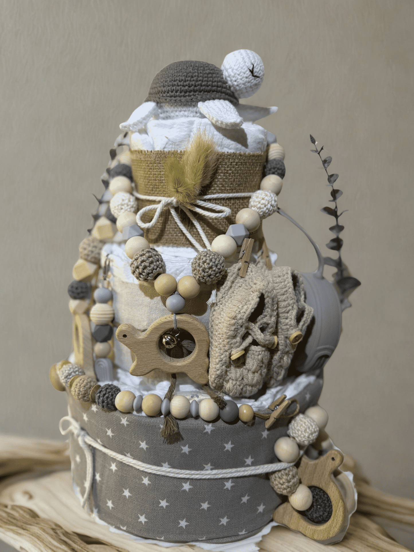 Neutral teddy bear diaper cake baby shower centerpiece featuring three tiers of rolled white diapers wrapped in natural burlap and soft gray star-print fabric. Top tier decorated with a crocheted teddy bear plush wearing a matching hat, accented with dried bunny tail grass and rustic twine bow. Adorned with silicone and crochet beaded pacifier clips in beige, gray, and cream tones, wooden bead garlands, and natural wood teething rings including animal-shaped designs. Includes a soft knitted baby bonnet and 