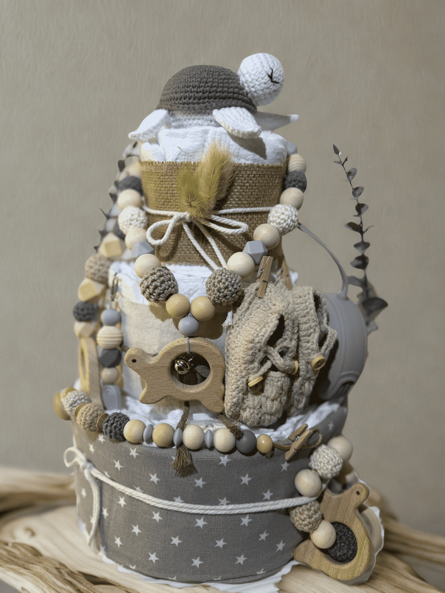 Neutral teddy bear diaper cake baby shower centerpiece featuring three tiers of rolled white diapers wrapped in natural burlap and soft gray star-print fabric. Top tier decorated with a crocheted teddy bear plush wearing a matching hat, accented with dried bunny tail grass and rustic twine bow. Adorned with silicone and crochet beaded pacifier clips in beige, gray, and cream tones, wooden bead garlands, and natural wood teething rings including animal-shaped designs. Includes a soft knitted baby bonnet and 