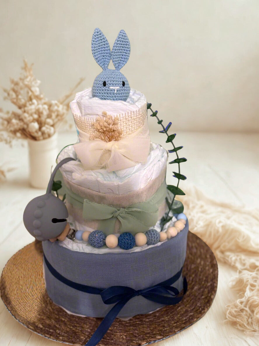 Blue and sage bunny diaper cake baby shower centerpiece featuring three tiers of rolled white diapers in soft, calming tones. The base tier is wrapped in dusty blue fabric and finished with a deep navy satin ribbon bow, creating a classic and elegant foundation.

The middle tier is accented with a muted sage green chiffon bow and coordinating trim, complemented by a silicone and crochet beaded pacifier clip in shades of navy, sky blue, cream, and natural wood. A matching gray pacifier case is attached to th